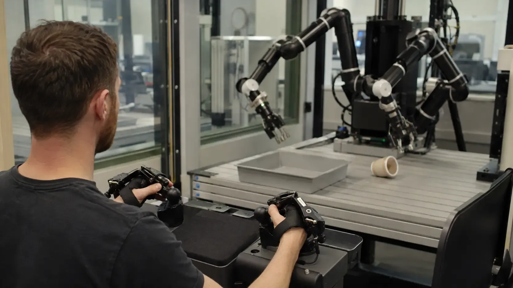 An operator at a teleoperation console watching two robot arms over a test table, a cup lying on its side beside the tray.