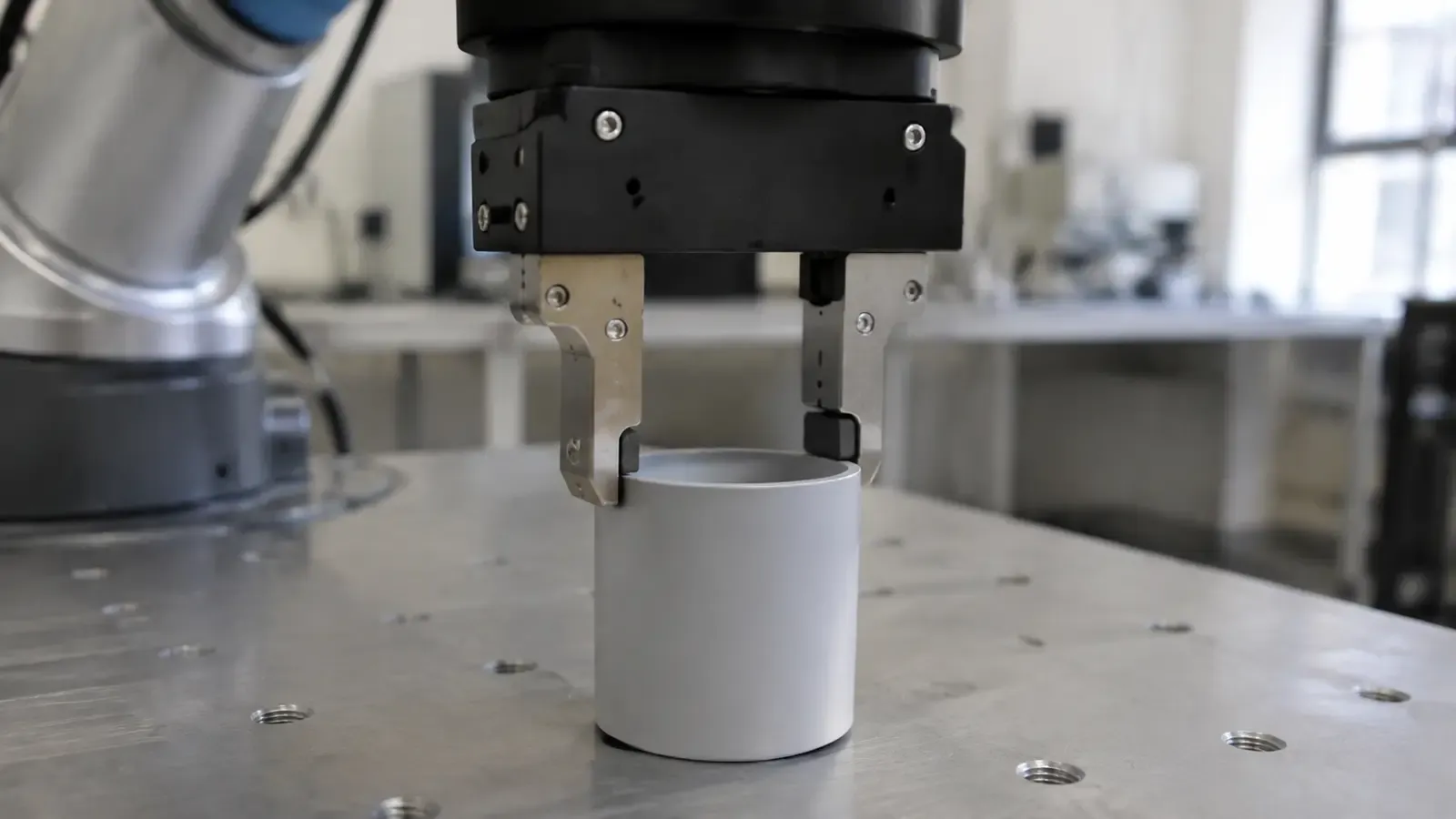 A two-fingered robot gripper lowered around a white cylinder standing on a steel test table.