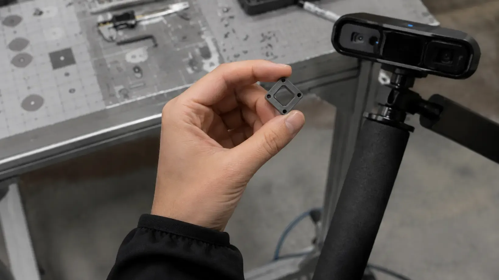A hand holding a small machined bracket up to a depth camera over a workbench laid out with parts.