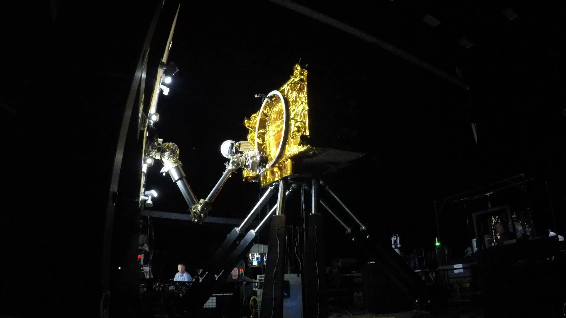 A gold-foiled satellite on a test rig, two robot arms aligned against it under studio lights.