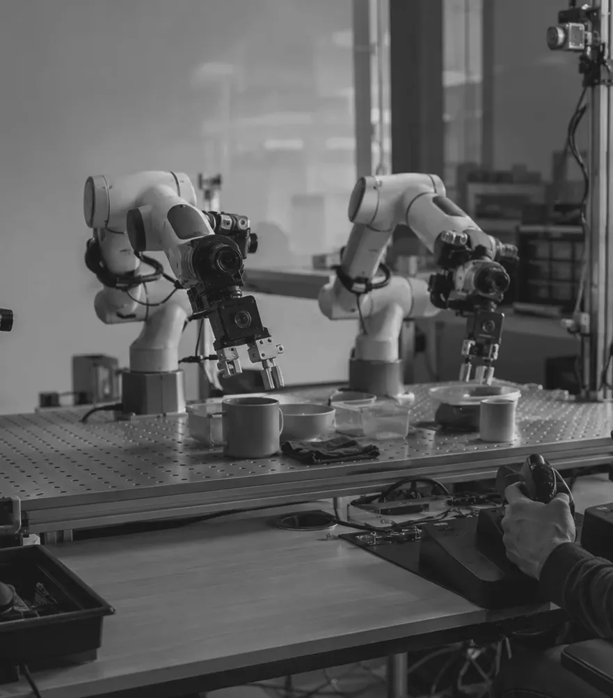 An operator driving a pair of robot arms across a workbench while overhead cameras record the run.