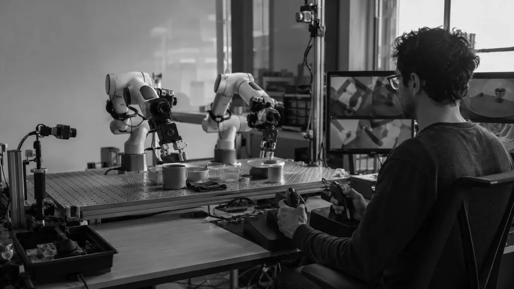 An operator at a workstation driving a pair of robot arms over a bench of small objects.